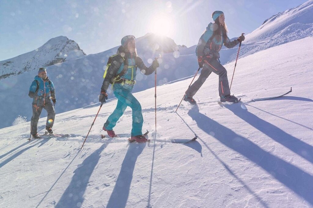 Three skiers with full gear ascending a snowy slope in bright sunlight, emphasizing safety in ski touring.