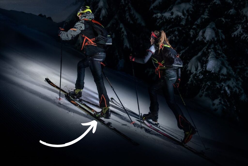 Night ski touring with headlamps, ascending a snowy mountain slope.  The white arrow indicates their upward climb.